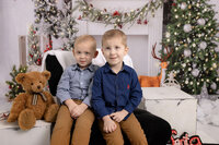 Two young children sit on a black couch surrounded by Christmas decorations, including a teddy bear, a Christmas tree, and festive garlands. Wearing blue shirts and brown pants, they smile at the camera in a cozy holiday setting captured by Jacksonville Cake Smash Photography.