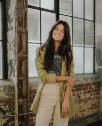 A woman with long dark hair stands smiling, dressed in a green jacket, grey shirt, and beige pants, in front of large industrial-style windows in a rustic setting, reflecting her expertise in branding and website design packages.