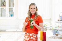 Phillipa Leseberg in an orange top holds a green juice bottle in her bright kitchen, surrounded by red and yellow alternatives. Her journey into alternative health reads like a memoir, filled with vibrant flavors and colorful experiments.