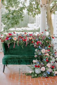 Gorgeous Wisconsin winter wedding photoshoot backdrop with vibrant winter-themed red, blue and pink flowers cascading on the back of and down to the floor of an emerald green velvet vintage love seat/settee couch rented by Beautifully Layered Event Rentals on a red brick herringbone path outdoors