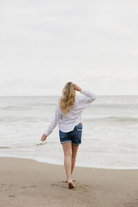 Brogan walking on the beach towards the water wearing a light white button up and jean shorts