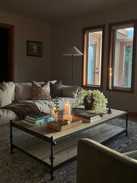 Bathroom with blue subway tile on the shower on the left. On the right is a single sink vanity in a neutral, beige paint with four drawers and an open shelf on the bottom. Above is mirror with a thin black frame and a silver sconces with a white shade on either side of the mirror.