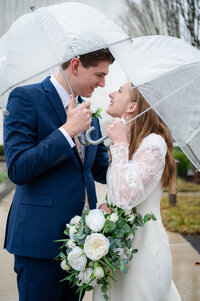 Bride and Groom hold umbrellas in the rain and smile at eachother