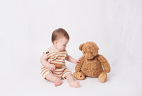 Little boy sitting with teddy bear wearing tan romper during cake smash photoshoot  in Brentwood Tennessee photography studio
