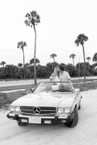 Couple shares a kiss in a vintage Mercedes 380SL convertible during their maternity shoot on St. Augustine Beach, surrounded by palm trees. Captured on a mix of 35mm film and digital.