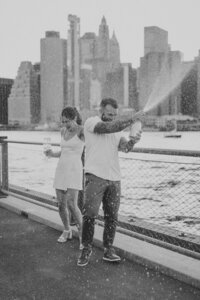 man and woman popping champagne while standing next to water with city in the background