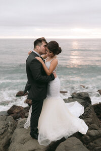 Bride and groom hug each other on rocks by ocean during sunset