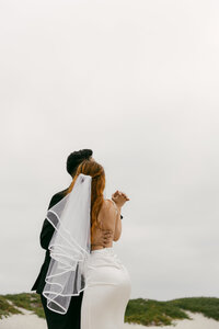 ride and groom facing away, standing close together on a cloudy beach with windswept veil and muted skies.