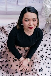 Woman sitting on a softly lit winter-themed set, wearing a black top and a light pink skirt with black floral patterns, smiling at the camera.