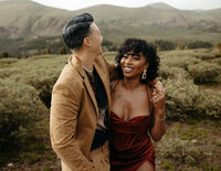A couple laughs together against a backdrop of rolling hills, the woman in a striking red dress and the man in a casual beige jacket