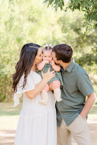 A couple stand on a sandy path at Katherine Fleischer Park. The mother holds their baby girl in between them. The parents kiss the baby on the cheeks, in this shot captured by an Austin Family Photographer