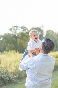 Smiling baby being lifted into the air by his dad during a sunny outdoor family photography session surrounded by green fields and trees in Connecticut.