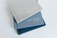 A layflat album with pink linen cover lies on a wood table, open  to reveal the thick pages and photos of engaged couple