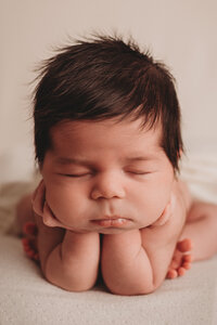 Newborn girl with a lot of brown hair sitting in a froggy pose, hands under chin and cheeks