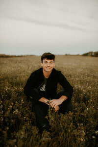 Photo of a senior boy sitting in a field of wildflowers smiling at the camera.