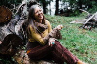 A woman with long brown hair wearing feather earrings sits cross legged on a log laughing at the camera. | Erica Swantek Photography
