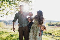 A father and children laughing together in a sunlit field as the mother leans in, with mountains and trees in the background.