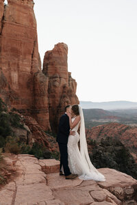 Bride and groom sharing a kiss on the red rock ledge during their Sedona elopement at Cathedral Rock with sweeping desert views in the background.