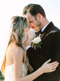 A bride and groom share a tender moment, embracing closely.  He wears a suit with a white rose boutonniere.