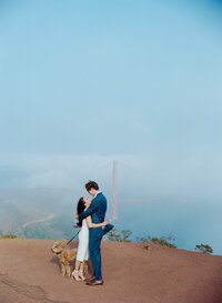 A happily engaged couple look passionately into each others' eyes in front of the Golden Gate Bridge as their brown dog in a leash looks on.