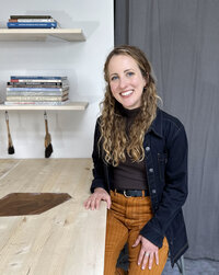 Master Printer Courtney Sennish smiling in a dark blue work coat and corduroy pants sits in a printmaking studio with art books and printmaking tools in the background.