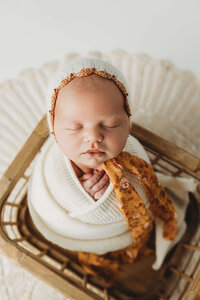 A peaceful baby sleeps soundly, swaddled in a rich maroon blanket, with a floral headband resting atop their head. The baby is cradled in a rustic wicker basket lined with soft fabric, creating a serene atmosphere.