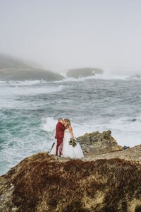 Newlyweds stand on cliffs overlooking crashing waves during an east coast adventure elopement. 