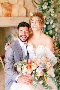 Bride and groom sitting together, laughing and holding a colorful wedding bouquet in front of a floral backdrop.