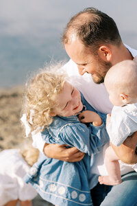 Dad holding baby boy and tickling daugher in blue dress. 