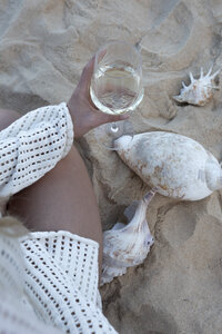 Glass of water and seashells next to woman’s hand at peaceful beach moment
