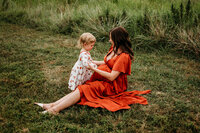 A pregnant mom is sitting on the ground wearing a flowy long dress and looking at a toddler who is touching her belly. They're sitting in a field with tall grass behind them.
