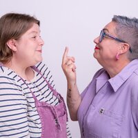 A picture of two humans on a lilac background. Lucy on the left is wearing dungarees and a striped t-shirt and has shoulder length brown hair. Pippa on the right is wearing a lilac jumpsuit, has a close cropped salt and pepper hair. Pippa is giving Lucy the bird, both are grinning.