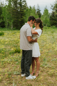 A man holding his fiance by the waist, facing slightly away from the camera so you don't see his face. His fiance has her hands around his neck and is smiling. Their foreheads are touching and there is trees in the background.