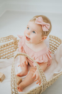 Baby girl in pink romper and bow smiles looking up with blue eyes sitting in moses basket during photo session
