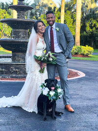 a bride and groom (groom in grey suite with a red tie) standing behind a black dog with a wedding flower collar on. Behind them is a fountaint.