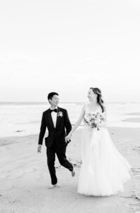 Barefoot bride and groom walk hand-in-hand along the shoreline at Hilton Head Island—captured in timeless black and white by luxury wedding photographer Amia Marcell.