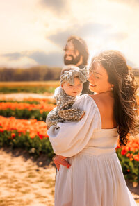 Toddler stands at the end of a long twisting lane that curves behind her. There's snow on the ground and tall brown grass behind her with trees in the background. She is wearing a tan coat and white snow hat with a pompom and is smiling big at the camera in this family photoshoot.