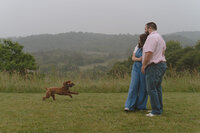 A photo of a husband and wife standing together cuddling in their back yard watching their dog run and play while it is raining. There are mountains in their background. Lifestyle couples photographer in Kingsport, TN. 