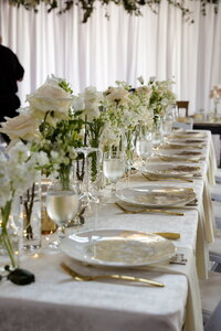 A beautifully set wedding table with white plates, gold cutlery, and crystal glasses features elegant white flower arrangements. The softly lit background with white drapes enhances the intimate atmosphere, akin to the refined setting found at Halcyon Watson’s exclusive venue.