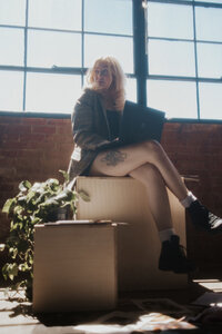 A woman is sitting on crates, in front of a large window, and typing on her laptop