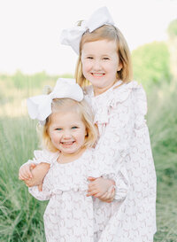 Two young sisters with matching white bows and pastel dresses smile brightly while embracing in a natural outdoor setting. Their joyful expressions and sweet bond capture the warmth and innocence of childhood, reflecting the light and timeless style of a Birmingham family photographer