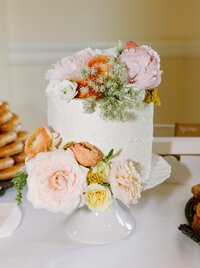 Small tall white wedding cake with large open roses, queen anne's lace on a white cake stand and a plate of doughnuts sitting behind the cake off to the left