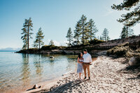 Bride and groom walking barefoot along the shoreline at Sand Harbor in Lake Tahoe, with pine trees, soft sand, and clear blue water in the background on a sunny day.