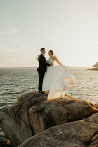 Bride and groom face each other and embrace at the top of a cliff by the water as the bride sways her wedding gown in the wind 