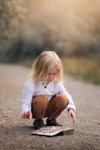 Little boy with long blonde hair crouched down to open a book on the ground.  Photo by Brittany Danielle.