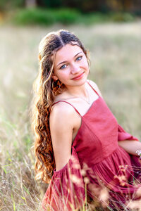 Stunning senior photos of girl in Central Florida posing near Lake Louisa State Park along the sandy lake shoreline. Senior girl is wearing red dress and crossing her arms while smiling at the camera.