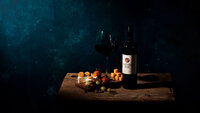 A moody and elegant image featuring a glass of red wine, a bottle of Matthew Fox wine, and a cheese board with assorted fruits and nuts. Photographed by Mica McCook, this composition captures the sophistication and richness of a wine and cheese pairing.