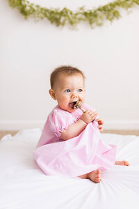 Baby girl in a pink dress sits and chews on an heirloom rattle