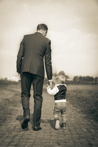 A tall man dressed in a suit walking down a cobblestone path while holding hands with a young boy.