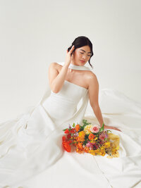 Film photo of a bride in a modern white gown with a structured scarf neckline, seated in a studio with a vibrant bouquet of poppies, roses, and orchids — captured by a film wedding photographer.